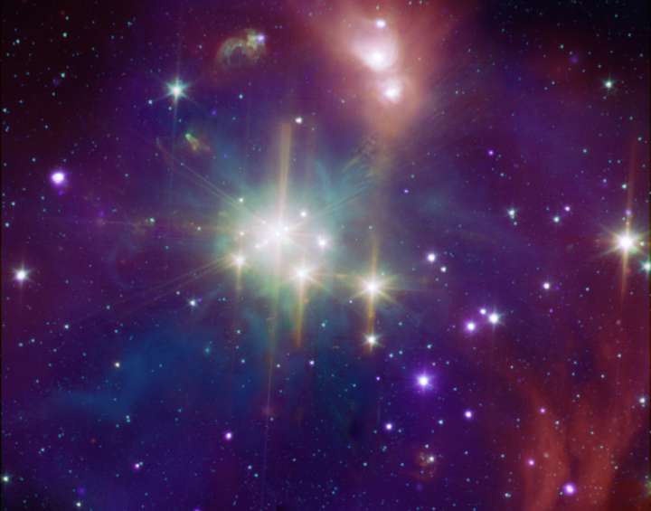 Die dicht gedrängten Sterne in der Bildmitte strahlen hell vor einem Hintergrund aus violetten und purpurfarbenen Wolken. Rundum sind weniger helle Sterne lose verteilt.