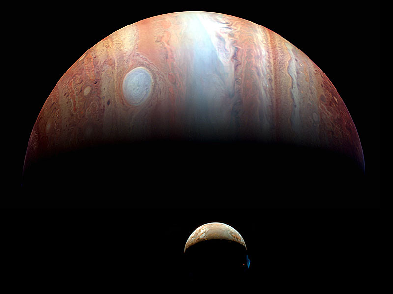 Vor dem halb beleuchteten Planeten Jupiter ist die Sichel des Mondes Io zu sehen, auf dem im Dunkeln heiße Lava leuchtet.