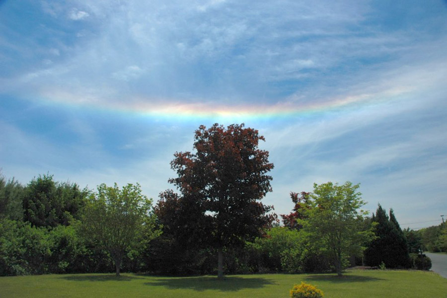 Am blauen, wolkigen Sommerhimmel leuchtet ein Regenbogen, der nach oben zeigt.