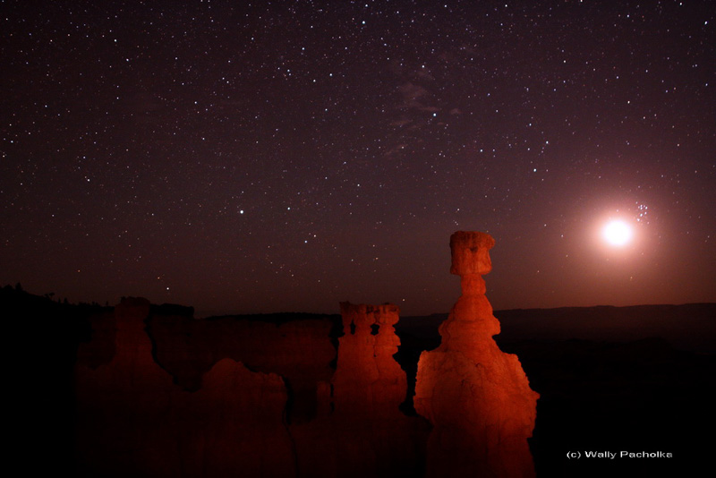 Rechts leuchten die Plejaden neben dem hellen Sichelmond, im Vordergrund stehen rechts nebn der Mitte rötliche Hoodoos, am Himmel sind einige Sternbilder erkennbar.