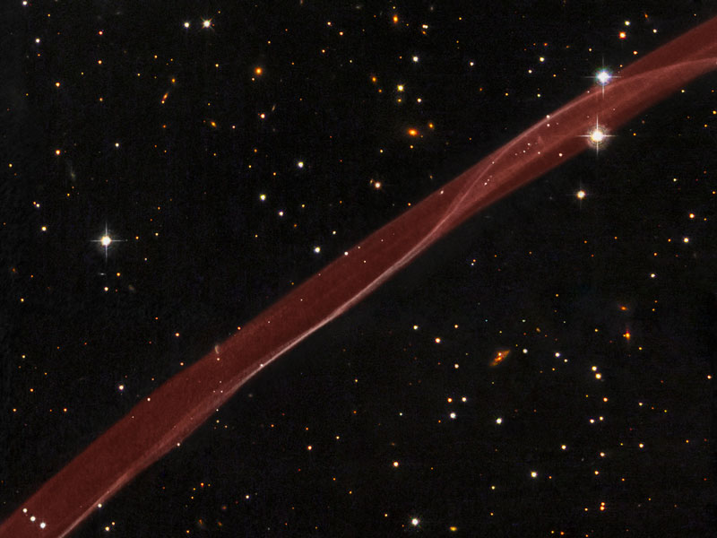Siehe Erklärung. Ein Klick auf das Bild lädt die höchstaufgelöste verfügbare Version. Ein rötliches Band verläuft diagonal von links unten nach rechts oben durch ein Sichtfeld mit einigen verstreuten Sternen.