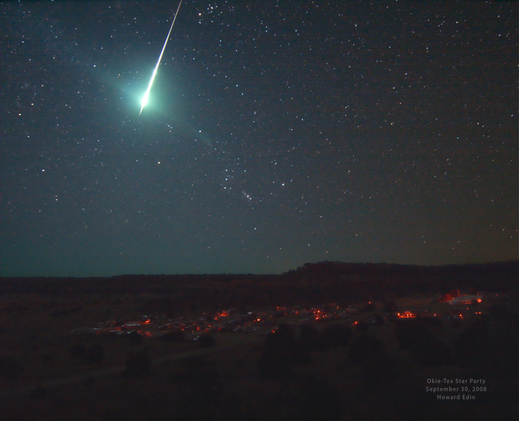 Am sternklaren Himmel blitzte eine Feuerkugel auf, am Sternenhimmel ist das Sternbild Orion zu sehen, unten ist die Silhouette einer Landschaft mit wenigen roten Lichtern.