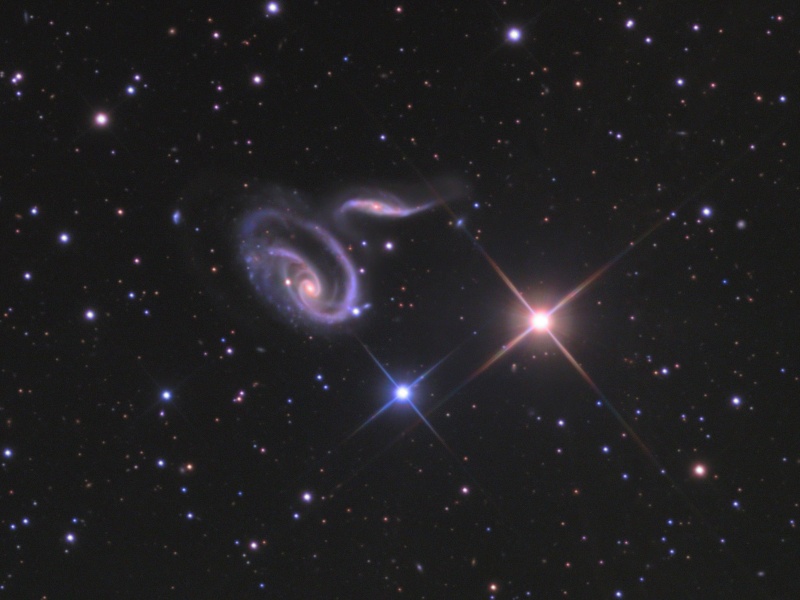Im Bild mit wenigen Sternen leuchten ein blauer und ein orangefarbener Stern und darüber zwei stark verzerrte Galaxien.