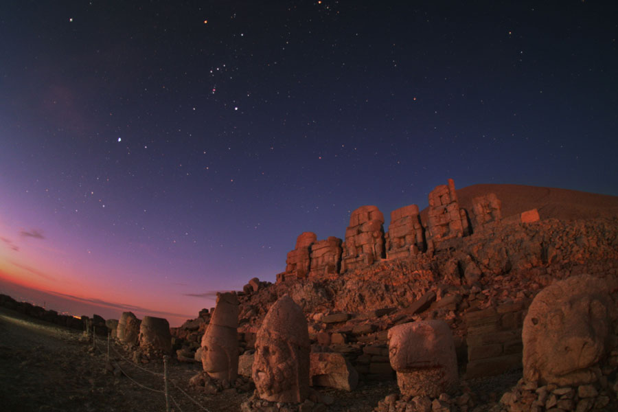 Hinter einer historischen Stätte mit gewaltigen Skulpturen leuchtet am Morgenhimmel in der Dämmerung das Sternbild Orion.