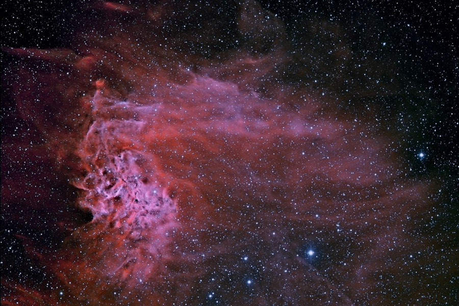 Im sternklaren Bild leuchtet ein rot-magentafarbenr Nebel, links ist ein dichtes wolkiges Gebilde, das nach rechts weht.