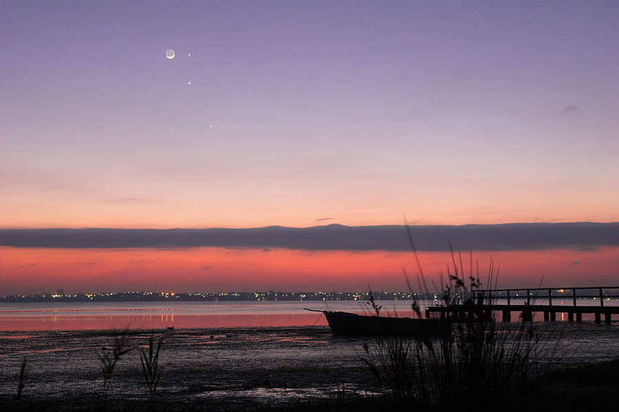 Am Himmel über einer stimmungsvollen Abendlandschaft mit See leuchtet eine Mondsichel und mehrere Planeten.