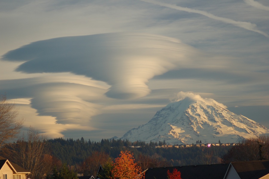 Hinter einer Waldlandschaft ragen schneebedeckte Berge hoch, darüber türmen sich linsenförmige Wolken auf, die aussehen wie UFOs.