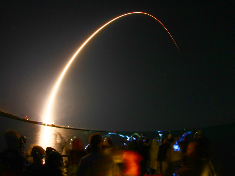 Am Nachthimmel zieht die leuchtende Abgas-Spur einer Rakete einen Bogen von links unten nach rechts oben.