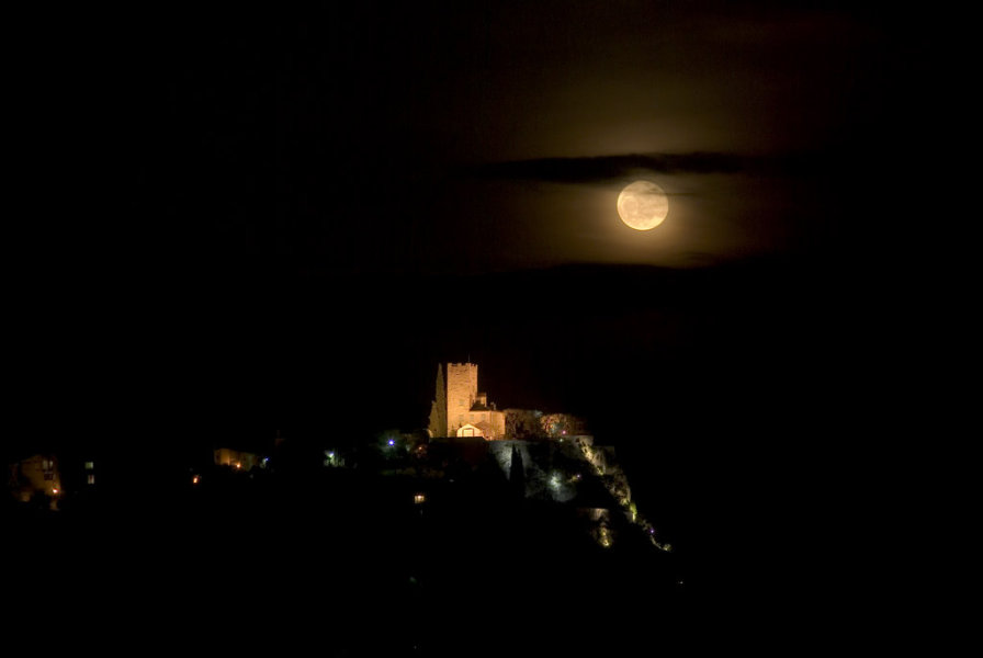 Über einer beleuchteten Burgruine auf einem Berg leuchtet der Vollmond, über dem ein Wolkenschleier liegt.