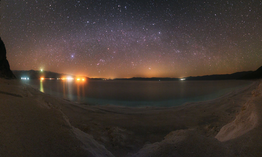 Der Blick reicht von einer Küste über Wasser, dahinter sind Lichter einer Beleuchtung, darüber wölbt sich ein Sternenhimmel.