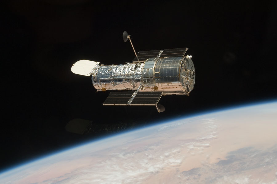 Das Weltraumteleskop Hubble schwebt über der Erde. Unten ist eine braune Landschaft mit weißen Wolken und dunklen Flecken zu sehen.