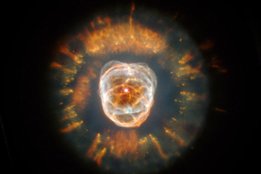 In der Mitte ist ein von hellen Wirbeln durchzogener runder Nebel, außen herum ist ein orangefarbener Ring aus nach außen gerichteten Strahlen angeordnet.