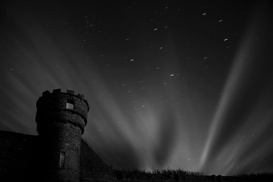 Hinter der Silhouette eines alten Turms sind seltsame strahlenartige Wolken und einiege Sterne zu sehen.