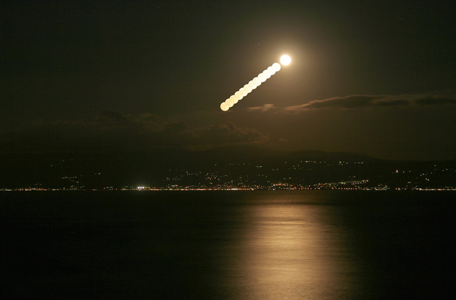 Der Mond geht über einer Wasserfläche auf, am hinteren Ufer liegt eine Stadt.