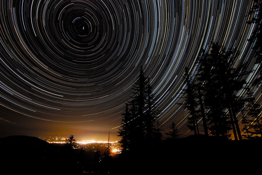 Das Bild zeigt Strichspuren an einem Nachthimmel hinter einem Nadelwald, im Hintergrund leuchten die Lichter von Grants Pass in Oregon.