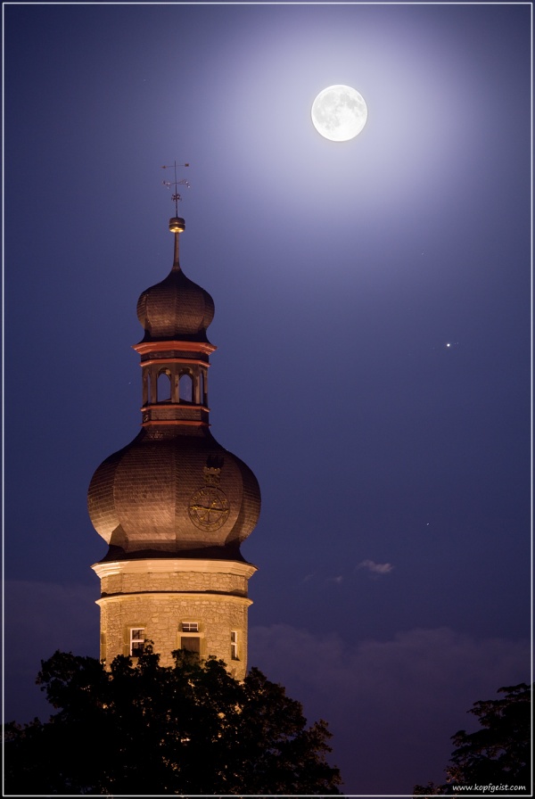 Über dem beleuchteten Kirchturm von Weikersheim, dessen Kuppel hinter einem Baum aufragt, leuchtet der Vollmond an einem dunkelblauen Himmel, rechts darunter ist der Planet Jupiter zu sehen.