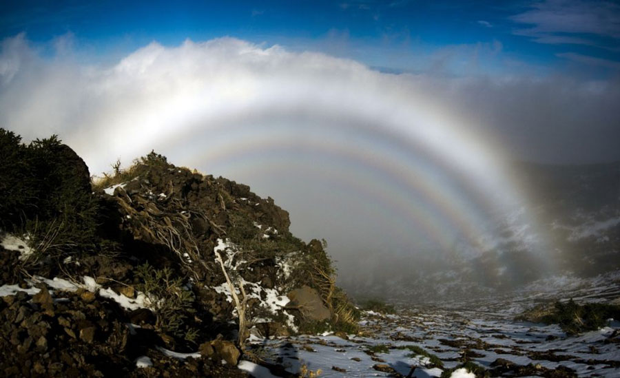 Von einer Berghöhe abwärts mit Felsen und etwas Schnee sind Wolken und Nebelschleier. Im Nebel sind konzentrische Nebelbögen zu sehen, eine Glorie, die eine Nebelhalle bildet.