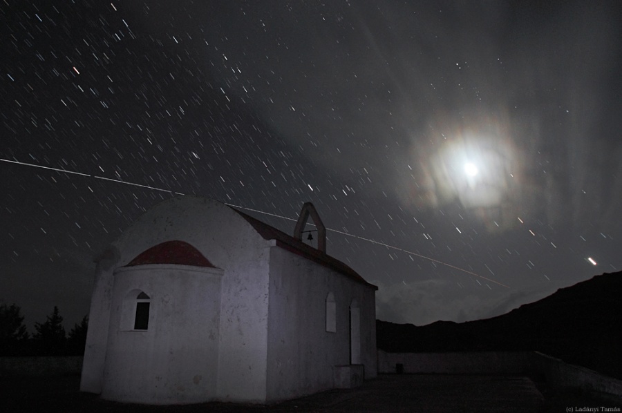 Hinter einer schwach beleuchteten Kapelle leuchtet der Mond, umgeben von einem Lichthof. Die Sterne am Himmel ziehen kurze Striche, die Raumfähre Atlantis zieht eine lange, waagrechte Leuchtspur.