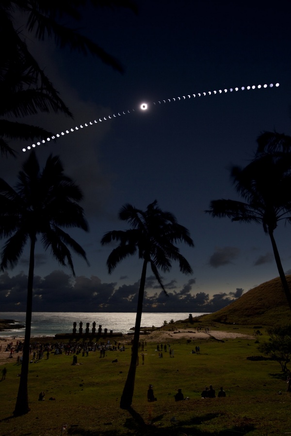 Die dunkle Szenerie blickt über einen Strand mit Palmen, am Ufer eines Gewässers stehen die Silhouetten von Moai. Am dunklen Himmel verlaufen Einzelbilder der totalen Sonnenfinsternis, in der Mitte leuchtet die Korona um den Erdmond.