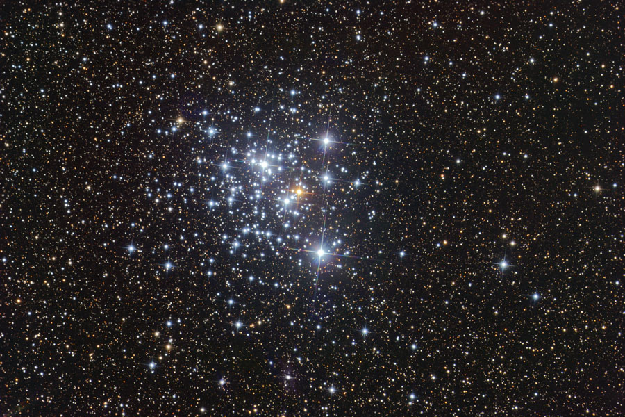 Wie ein Schmuckkästchen glitzern die Sterne eines offenen Sternhaufens inmitten von weniger hellen Sternen. In der Mitte leuchtet ein roter Stern, umgeben von blauen Sternen.