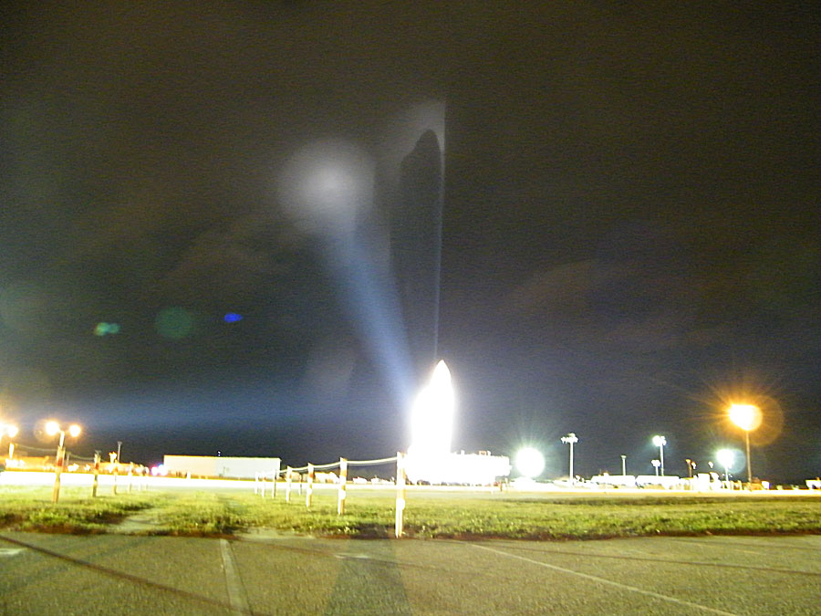 Hinter einem grün bewachsenen Gelände ragt eine weiße Gestalt auf und wirft ihren Schatten auf einen Nebel dahinter.