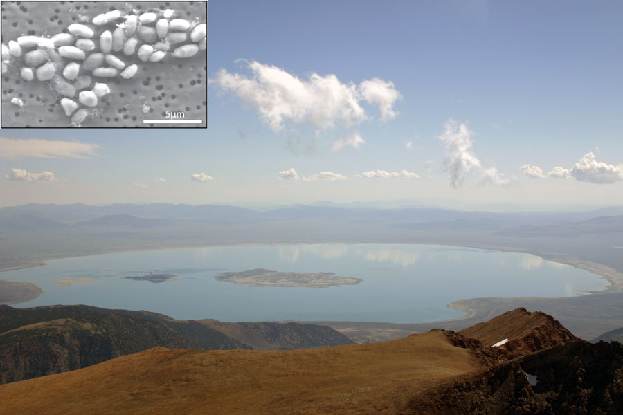 Von einem Berg aus ist unten ein runder See mit einer Insel in der Mitte zu sehen, dahinter reichen Berge bis zum leicht bewölkten Horizont. Links oben ist ein Einschub mit weißen ovalen Formen.