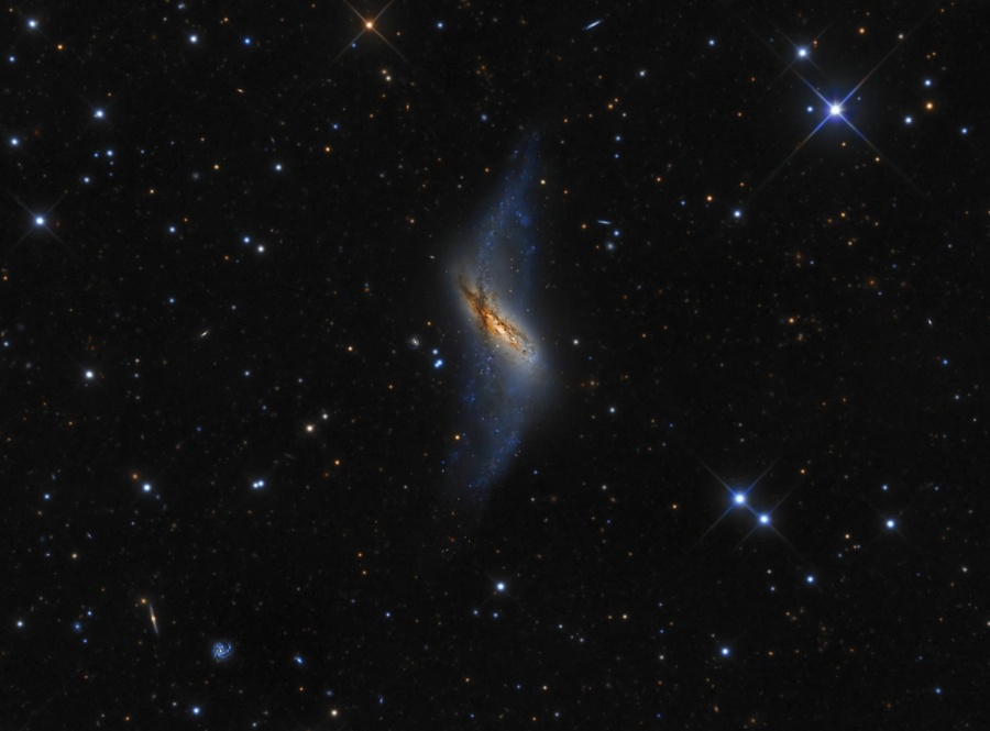 Mitten im Bild schwebt eine seitlich sichtbare, verkrmmte Polarringgalaxie, die von einem Ring aus Sterntrümmern gekreuzt wird. Darum sind Sterne lose verteilt.