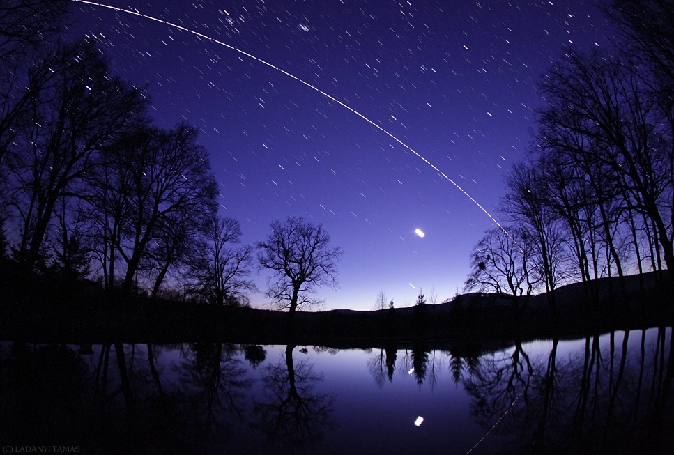 Über einem ruhigen See breitet sich ein dunkelblauer Himmel mit Strichspuren von Sternen aus, die sich im Wasser spiegeln. Hinten am Ufer stehen die Silhouetten von nackten Bäumen. Am Himmel verläuft eine helle gekrümmte Spur von links oben nach rechts unten. das hellste Licht am Himmel ist der Mond.