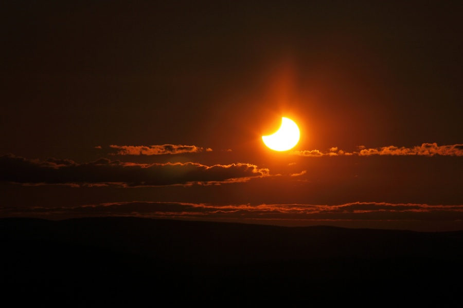 Über ein paar von hinten beleuchteten Wolken leuchtet eine breite Sonnensichel am dunklen Himmel.