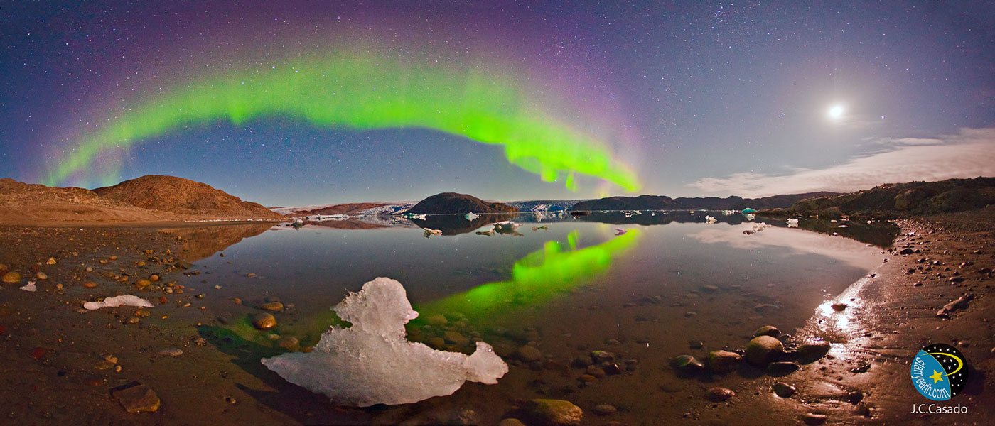 Über einem spiegelglatten Gewässer in einer felsigen Landschaft wölbt sich am klaren Himmel ein grünes Polarlicht, das oben purpurfarbene Ränder hat. Rechts leuchtet der Mond.