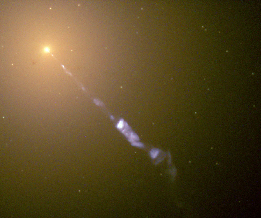 Links oben ist ein gelb leuchtendes Galaxienzentrum, von diesem strömt ein langer, blauer Strahl, der teilweise unterbrochen ist.
