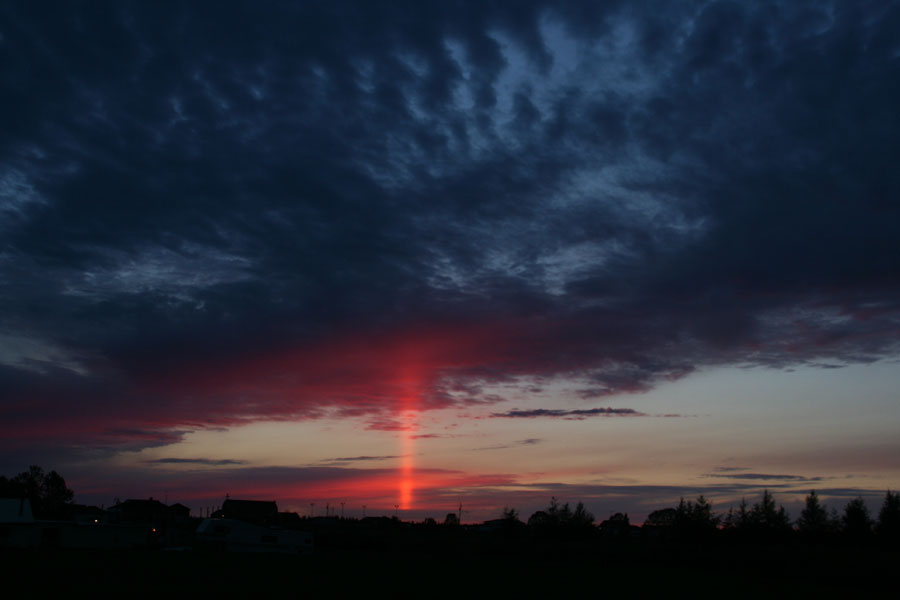 Unter dunklen Federwolken hoch am Himmel und über der dunklen Silhouette eines Waldes steigt eine leuchtendrote Lichtsäule auf.