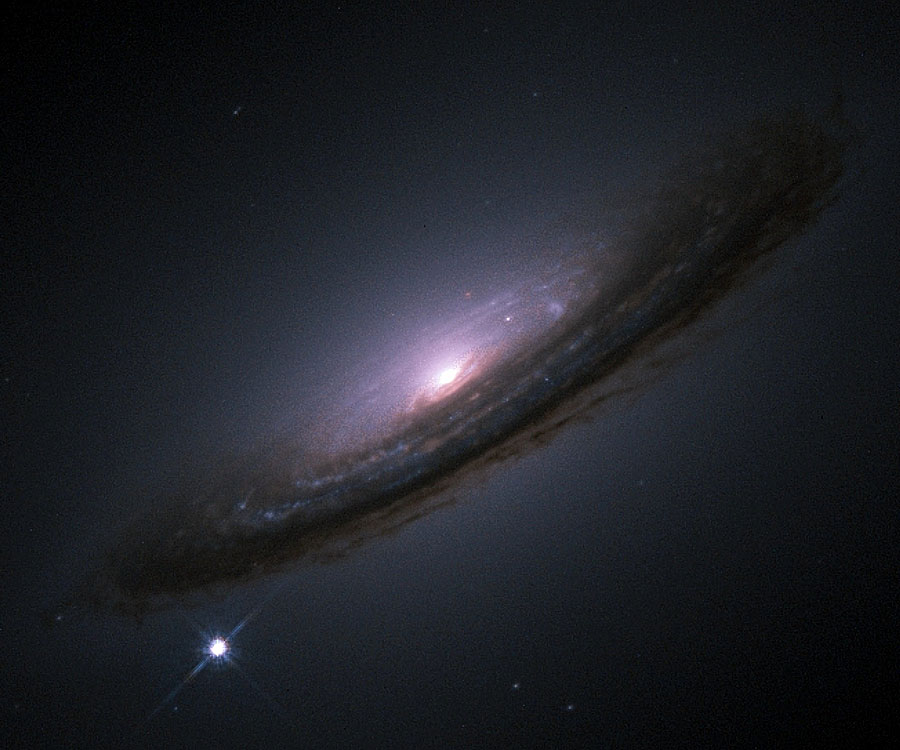 In der Mitte leuchtet ein helles Galaxienzentrum, das von viel Staub umgeben ist. Wir sehen die Galaxie schräg von der Seite. Vorne verläuft ein breiter Staubwulst, der die Galaxie vorne verdeckt.