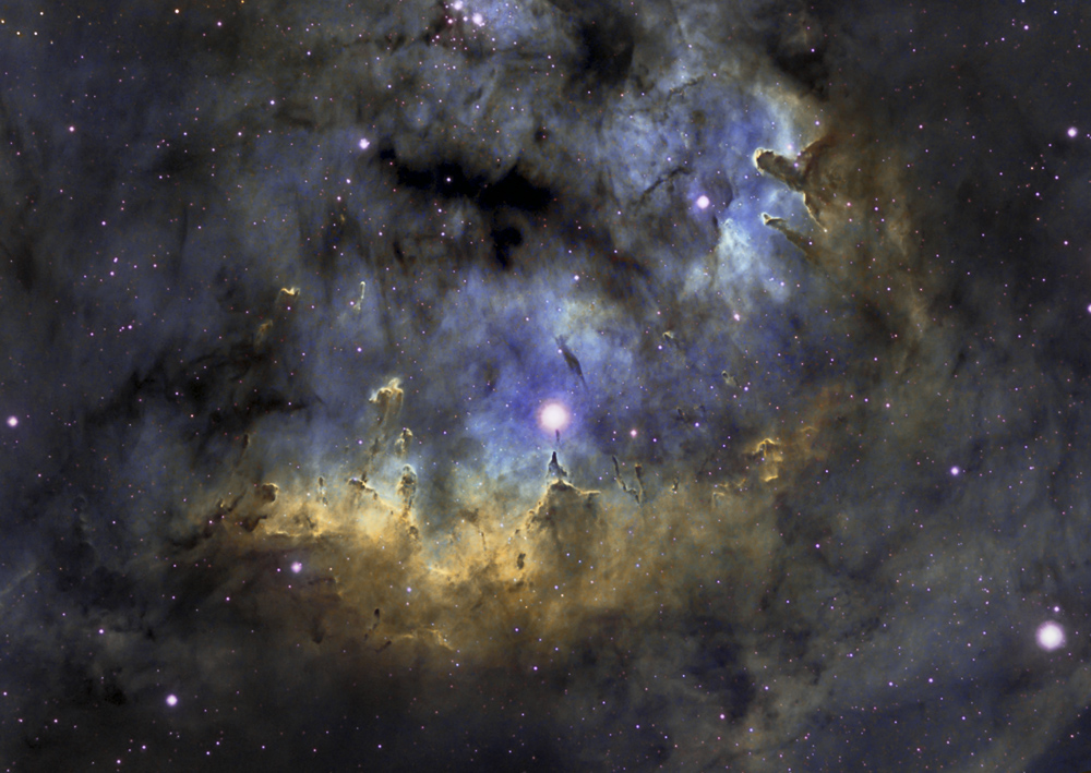 Hinter einem Gewirr aus dunklen Staubnebelfetzen leuchtet ein blauer Nebel im Hintergrund, unten ist ein ockerfarben beleuchtetes Staubnebelfeld.