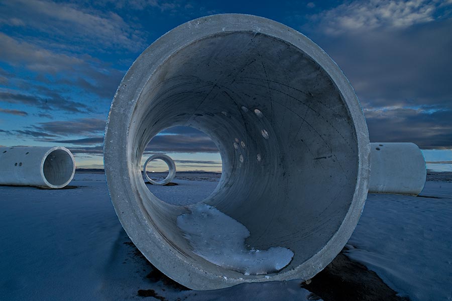 Hinter zwei Sonnentunneln geht die Sonne am Horizont unter. Die Landschaft ist blau und dämmrig, am blauen Himmel stehen viele Wolken. In der vorderen Betonröhre liegt Schnee, auch die Landschaft ist verschneit.