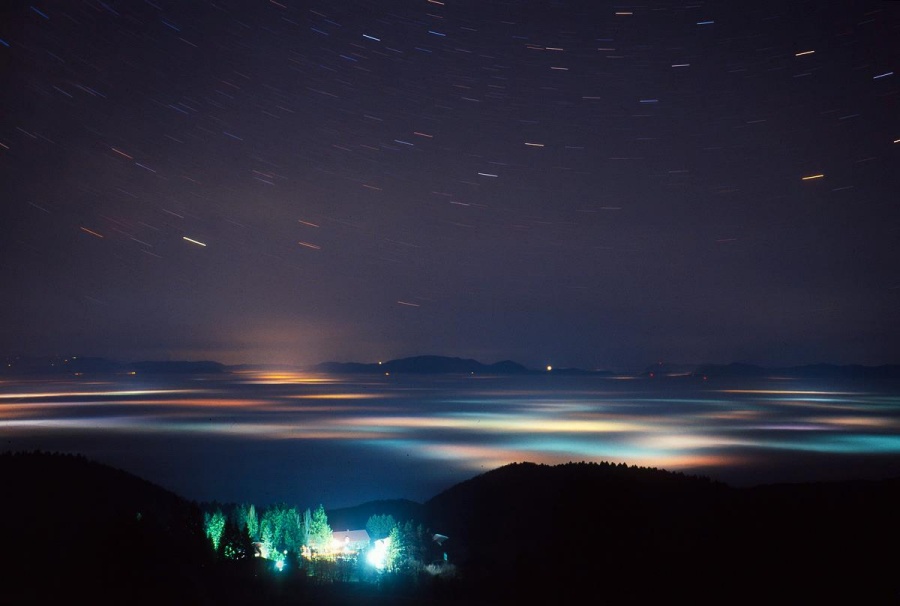 Die Aussicht zeigt ein beleuchtetes Gebäude mit Bäumen am unteren Bildrand, dahinter sind bunt beleuchtete Hochnebel über Zagreb in Kroatien. Am Himmel oben sind Strichspuren von Sternen.