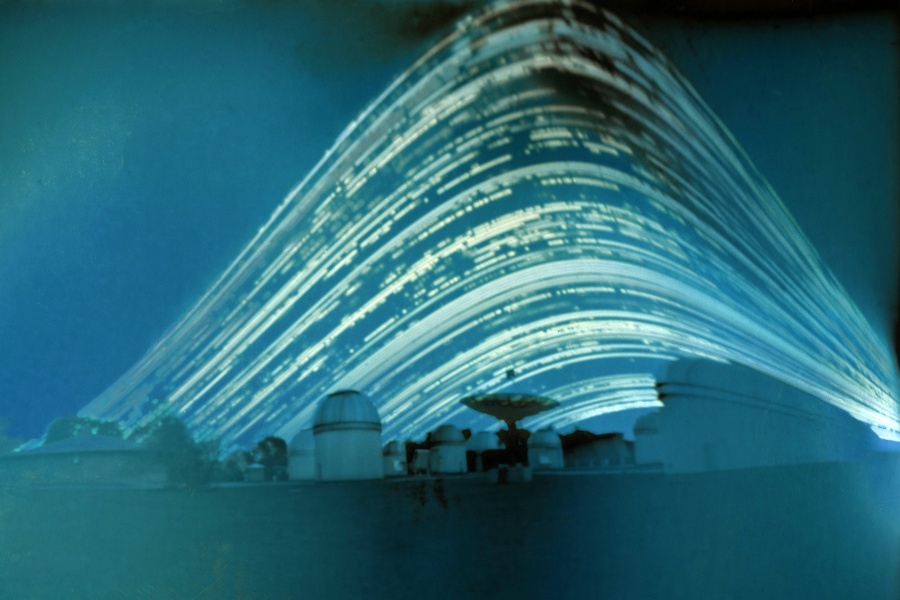 Das blaugrün gefärbte Bild zeigt ein Observatorium mit mehreren Teleskopkuppeln und einer Radioschüssel. Es wirkt etwas verzerrt. Darüber verlaufen helle Strichspuren in Schichten. Die Spuren sind gelegentlich lückenhaft.