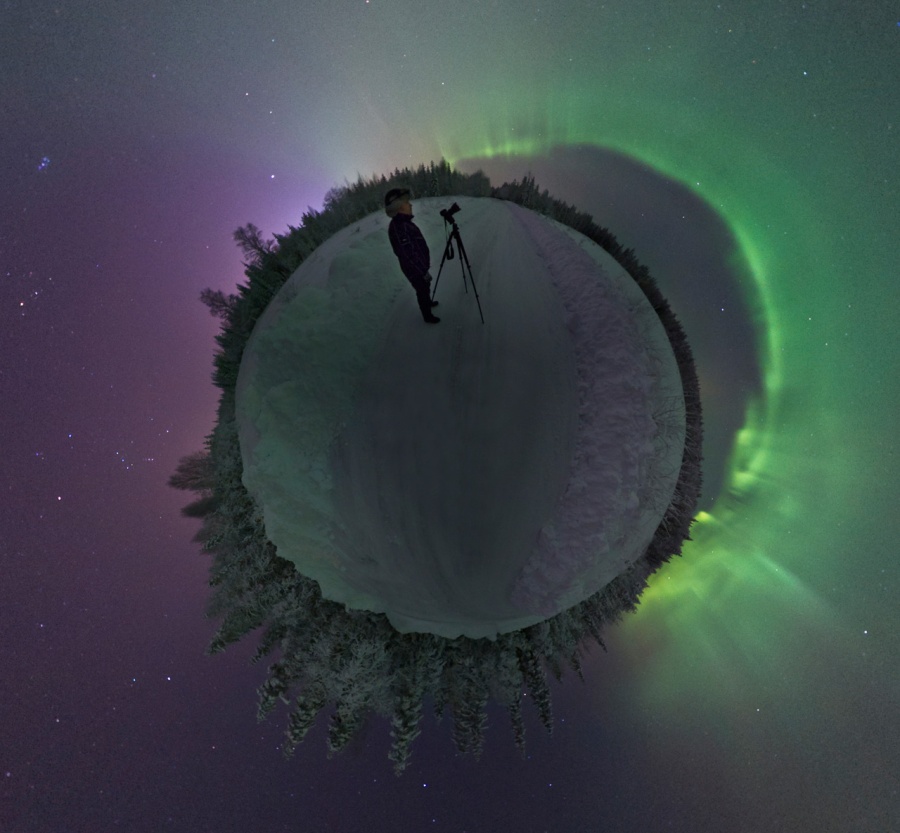 Ein kleiner Planet ist von Schnee bedeckt, es ist Nacht. Rechts leuchtet ein helles grünes Polarlicht in einem Bogen, links ist der Himmel lila. Am Horizont ist Wald, oben steht ein Fotograf mit Kamera.