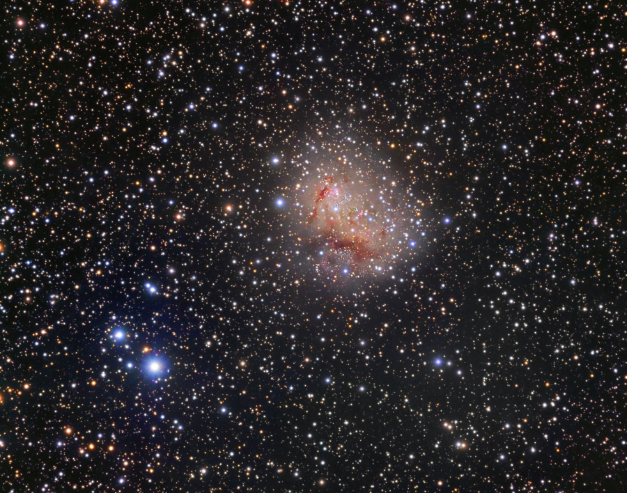 Hinter den Sternen und Staubschleiern der Milchstraße befindet sich die Zwerggalaxie IC 10, die hier abgebildet ist. Sie ist von rötlich-braunen Staubbahnen überzogen.