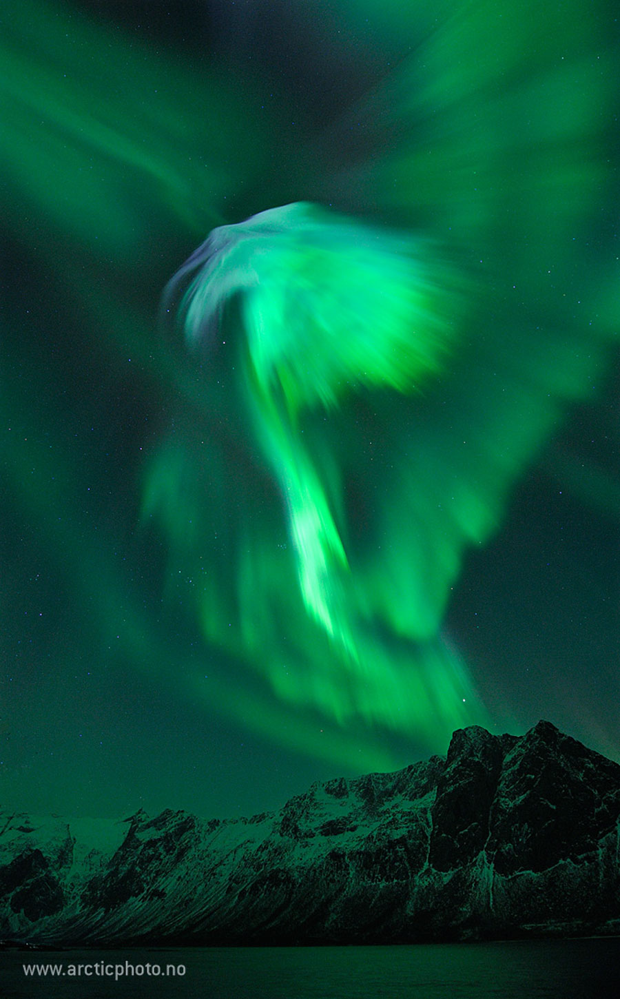 Am Himmel breitet sich ein grünes Polarlicht aus. Unten sind teilweise verschneite Berge von dem grünen Licht beleuchtet.