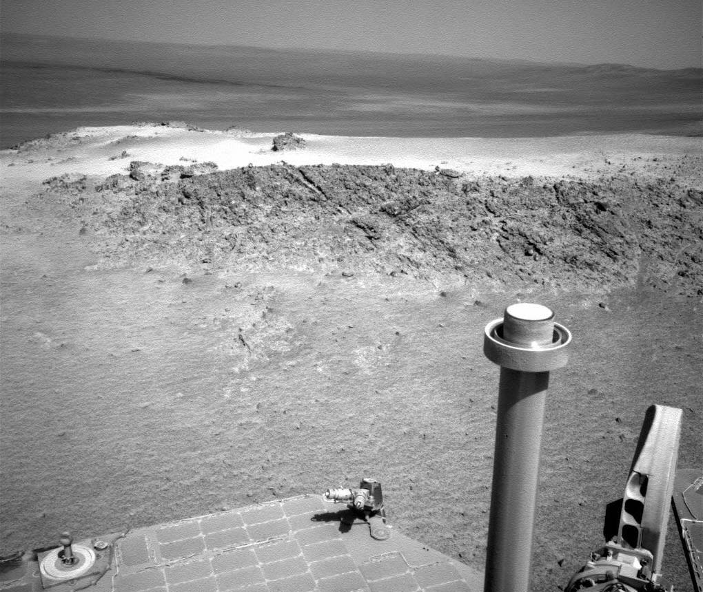 Der Rover Opportunity blickt über Solarpaneele und einen Mast im Vordergrund auf eine Marslandschaft mit einem Felshang im Vordergrund.