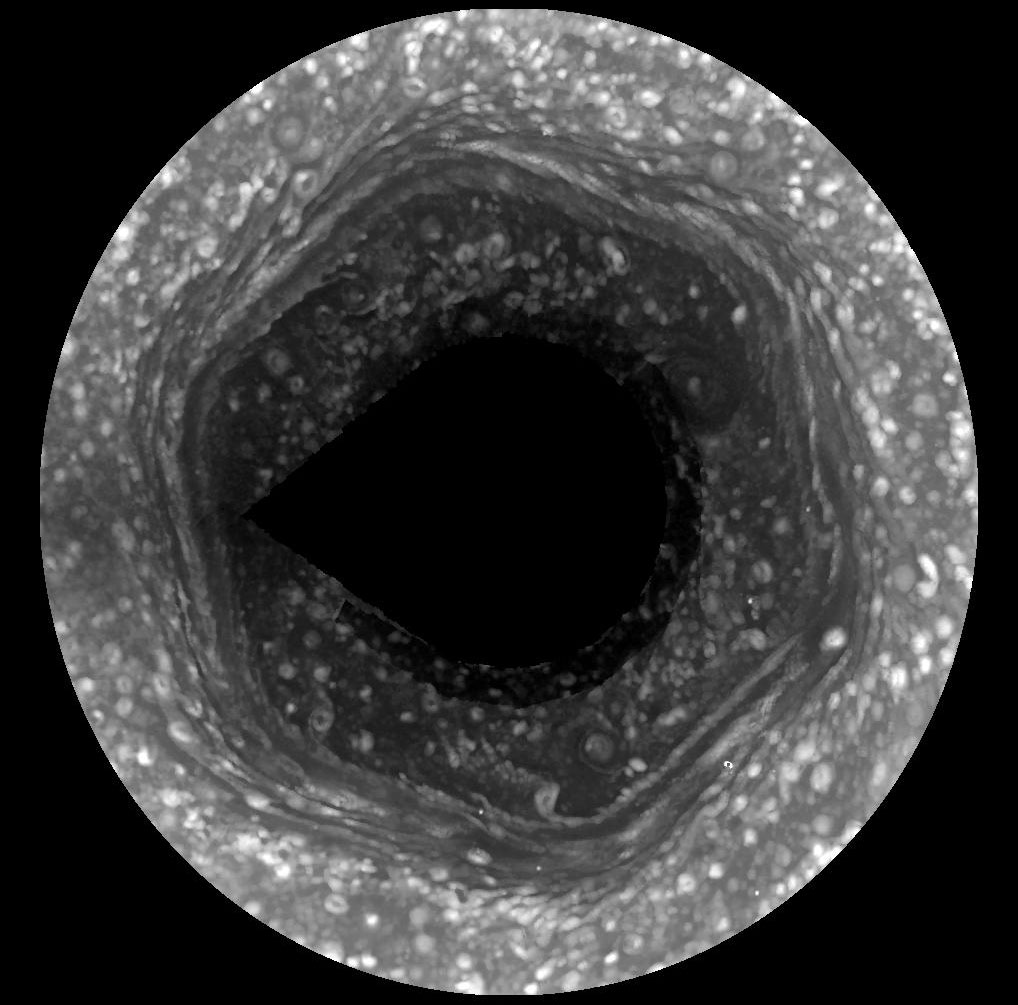 Im Bild ist ein runter Ausschnitt von Saturns Nordpol. In der Mitte ist eine dunkle runde Struktur, um diese herumverläuft ein sechseckiger Nebel. Die Wolken sind von weißen Flecken übersät.
