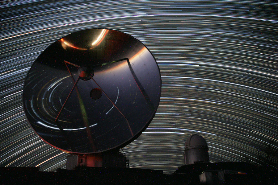 Hinter einem Radiotelesiop, in dem sich der Vordergund spiegelt, der hinter der Kamera liegt, und einer Teleskopkuppel zienen Sterne ihre Strichspuren am Nachthimmel.
