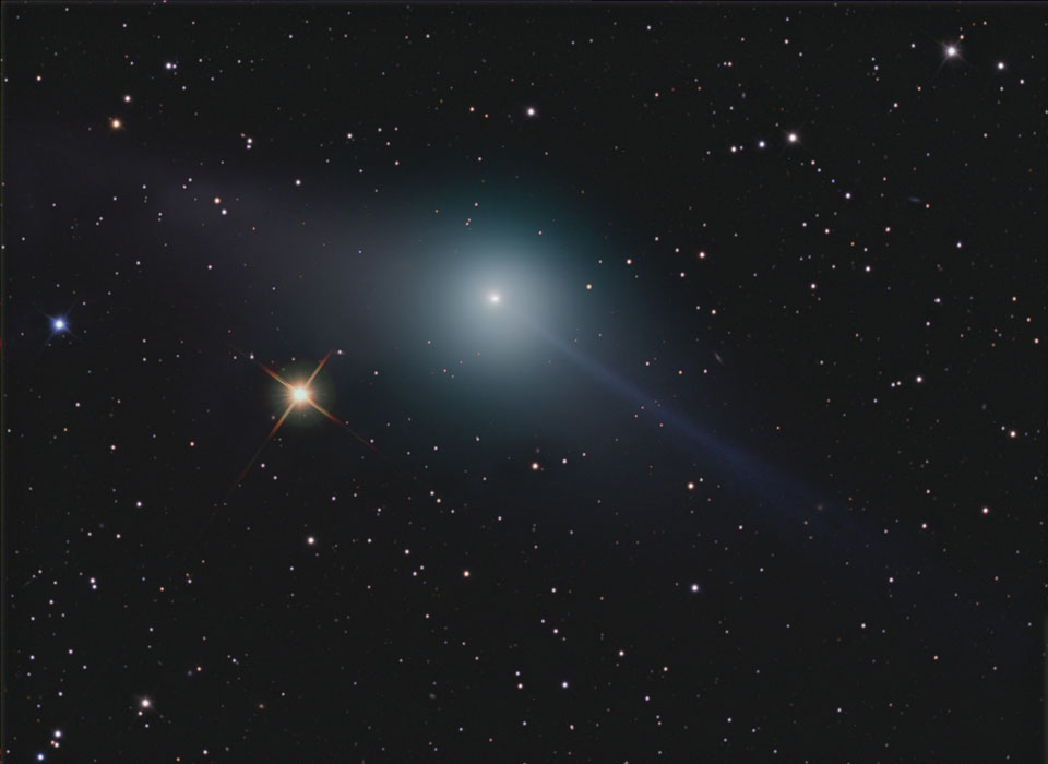 Der Komet Garradd im Bild besitzt scheinbar zwei Schweife, er ist von einer weißlich-grünen Koma umhüllt. Links daneben leuchtet ein gelber, gezackter Stern. Im Hintergrund sind viele kleine Sterne verteilt.