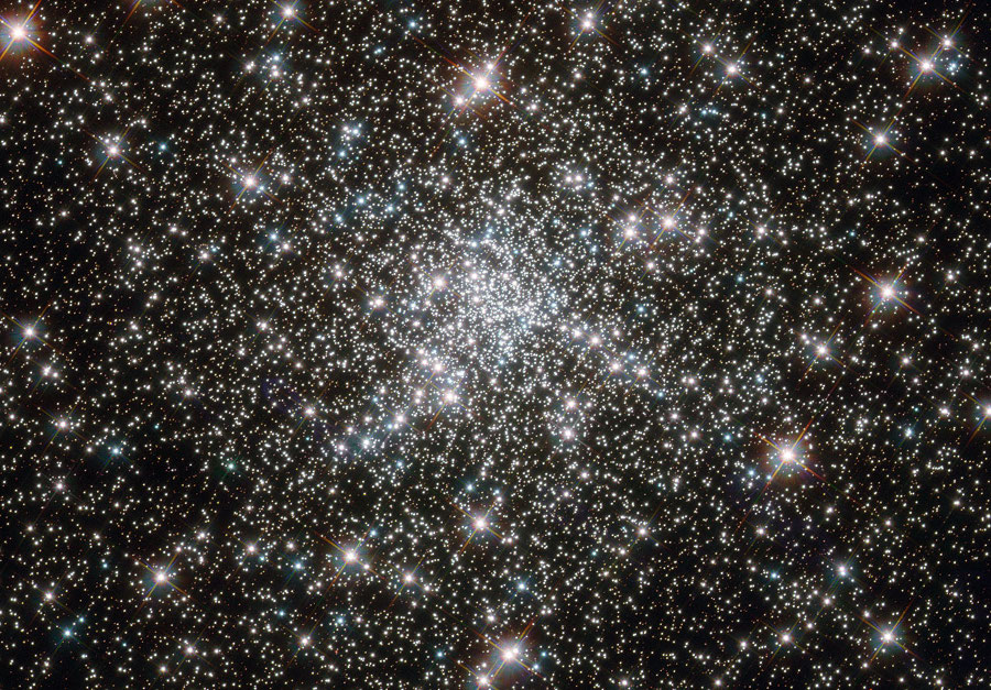 Mitten im Bild ist ein Kugelsternhaufen mit sehr vielen Sternen, eine größere Anzahl leuchtet hell hervor.