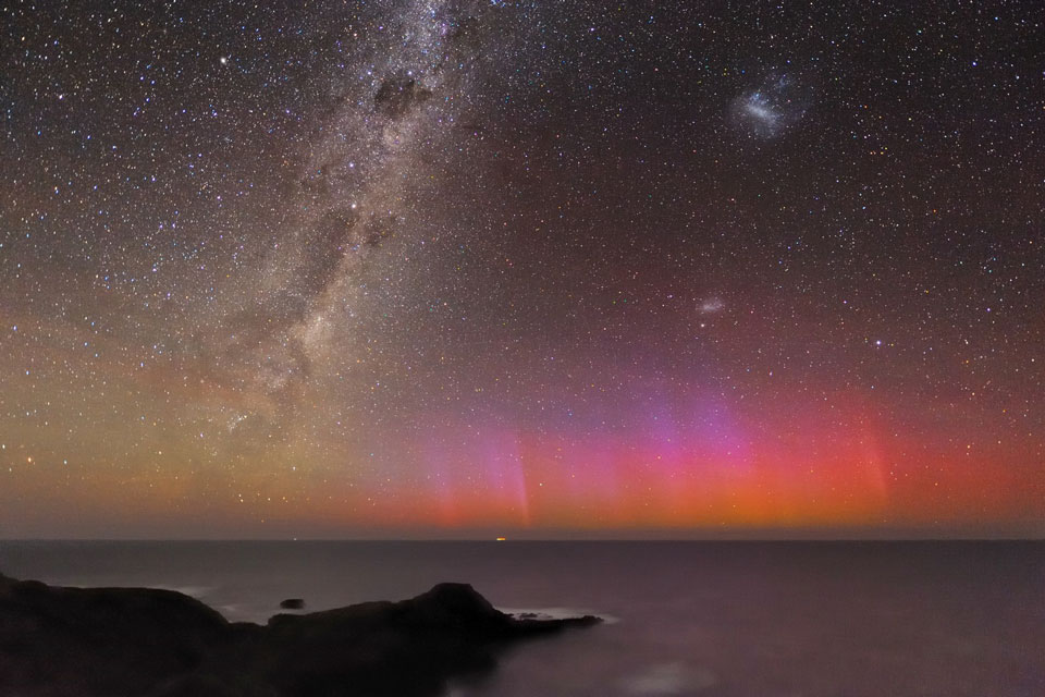 Über dem Horizont des Meeres leuchten rote Polarlichter, am Himmel stehen zahlreiche bekannte Objekte wie die Milchstraße und die Magellanschen Wolken.