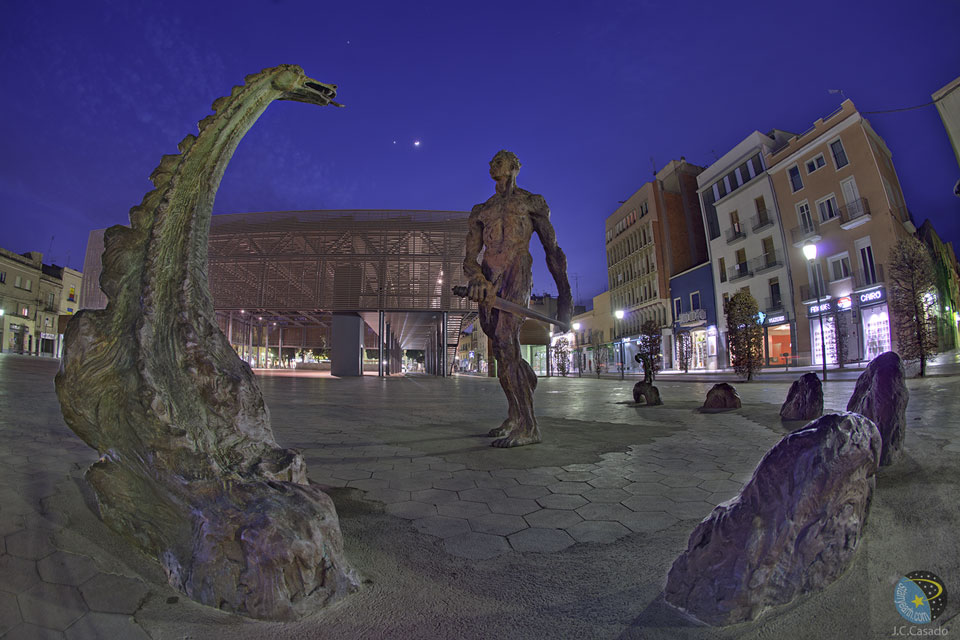 Hinter einem hell erleuchteten Platz mit einigen Statuen, die einen Drachen und einen Drachentöter darstellen, strahlen zwei Planeten am blauen Himmel.