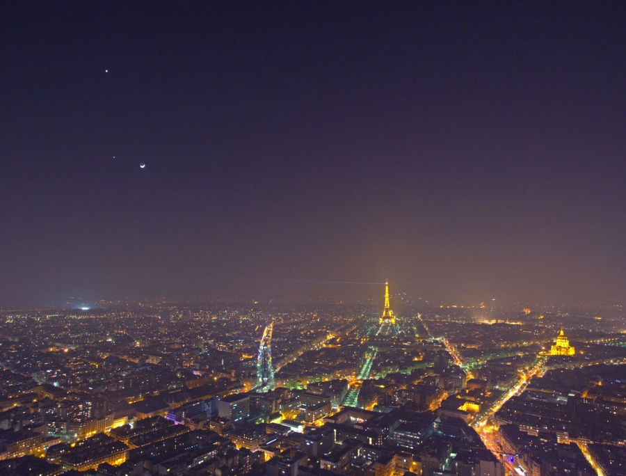 Mitten in einer riesigen, lichtverschmutzten Stadtlandschaft von Paris ragt der in hellem Gelb beleuchtete Eiffelturm heraus. Am Himmel leuchten Planeten und der Sichelmond.