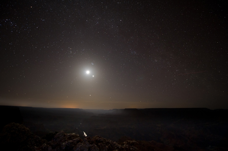 Am dunklen Nachthimmel leuchten über dem Horizont zwei Lichter, der Mond neben dem Planeten Venus. Unten windet sich der Colorado River durch die Schlucht des Grand Canyon.