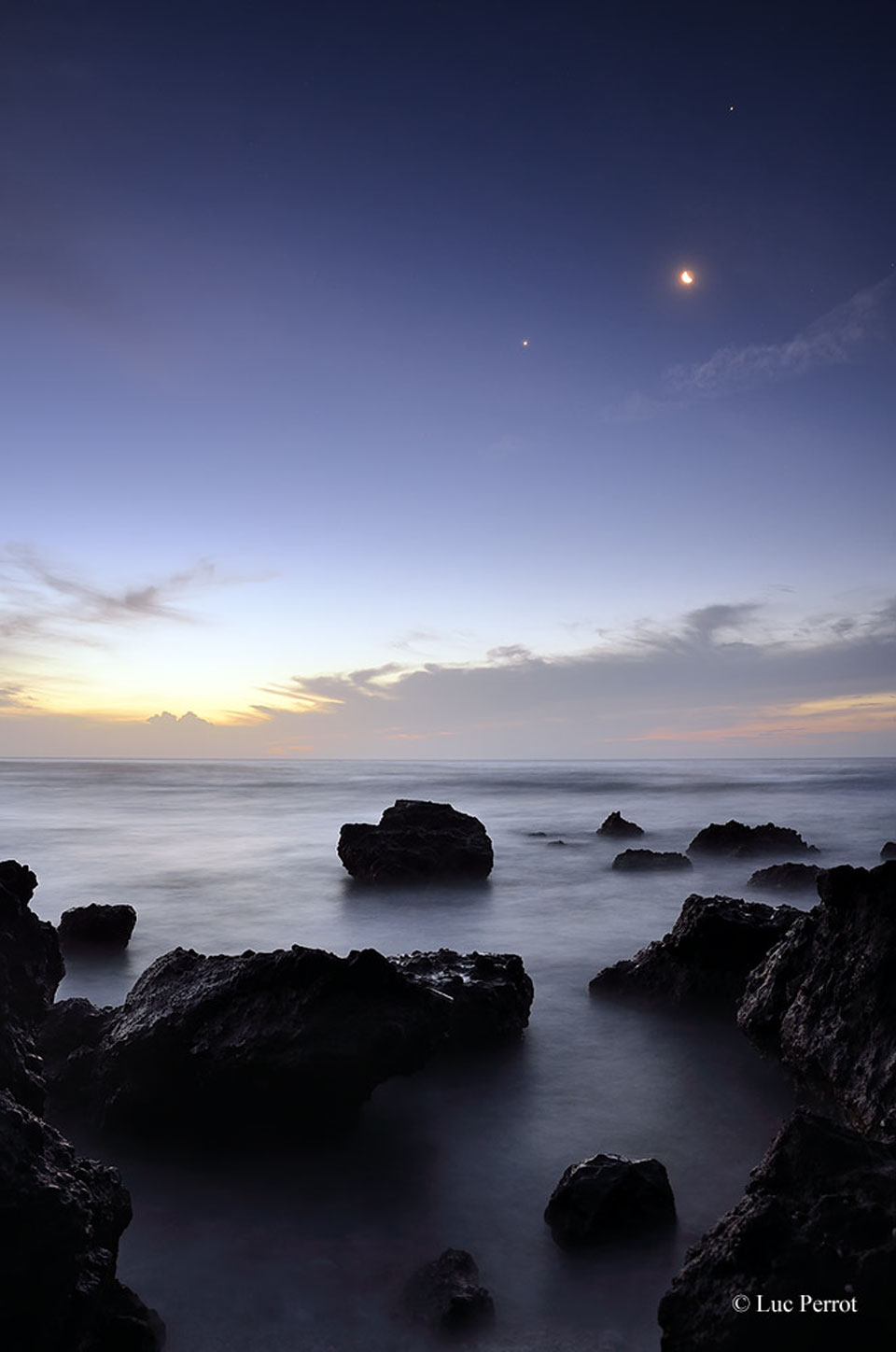 Über einer Küste mit riesigen Felsen stehen die Planeten Venus und Jupiter beim hellen Sichelmond.